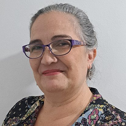 Woman with grey hair in a floral top and purple glasses smiling at the camera.
