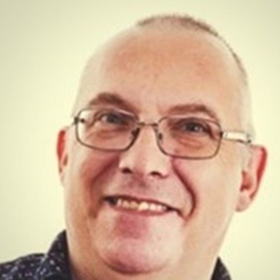 Smiling man with glasses and short hair, wearing a patterned shirt.