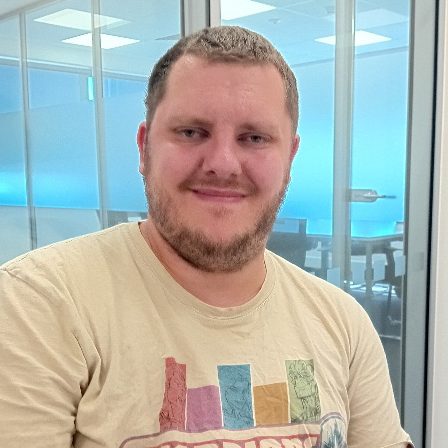Smiling man in a light-coloured t-shirt, sitting in a modern office environment.