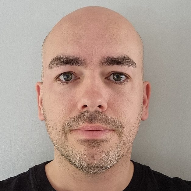 Male with a bald head and short facial hair, wearing a black shirt against a light background.