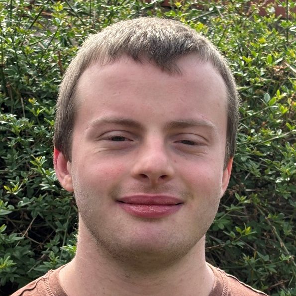 Young man with short hair, smiling, in front of greenery.