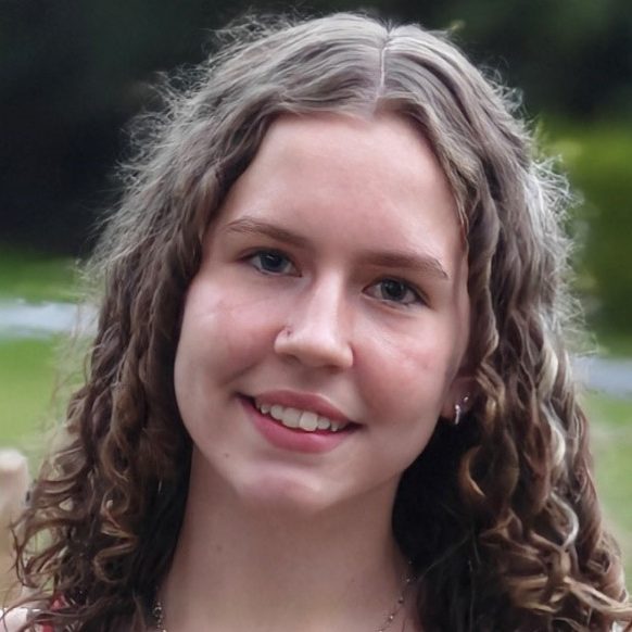 Ruby - ACC - Co-Trainer Young woman with curly hair smiling outdoors, wearing a red top against a natural background.