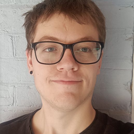 Young person with short brown hair, glasses, and a slight smile against a pale wall.