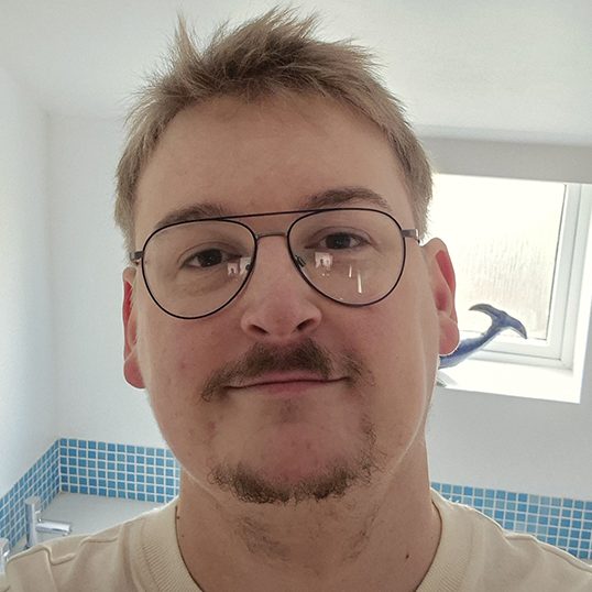 A person with short hair and glasses smiles in a bright, tiled bathroom.