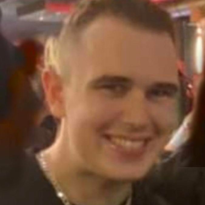 Smiling young man with short hair and a chain necklace, in a social setting.