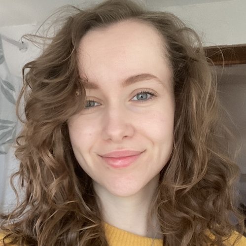 A woman with curly hair smiles, wearing a yellow sweater, in a bright indoor setting.