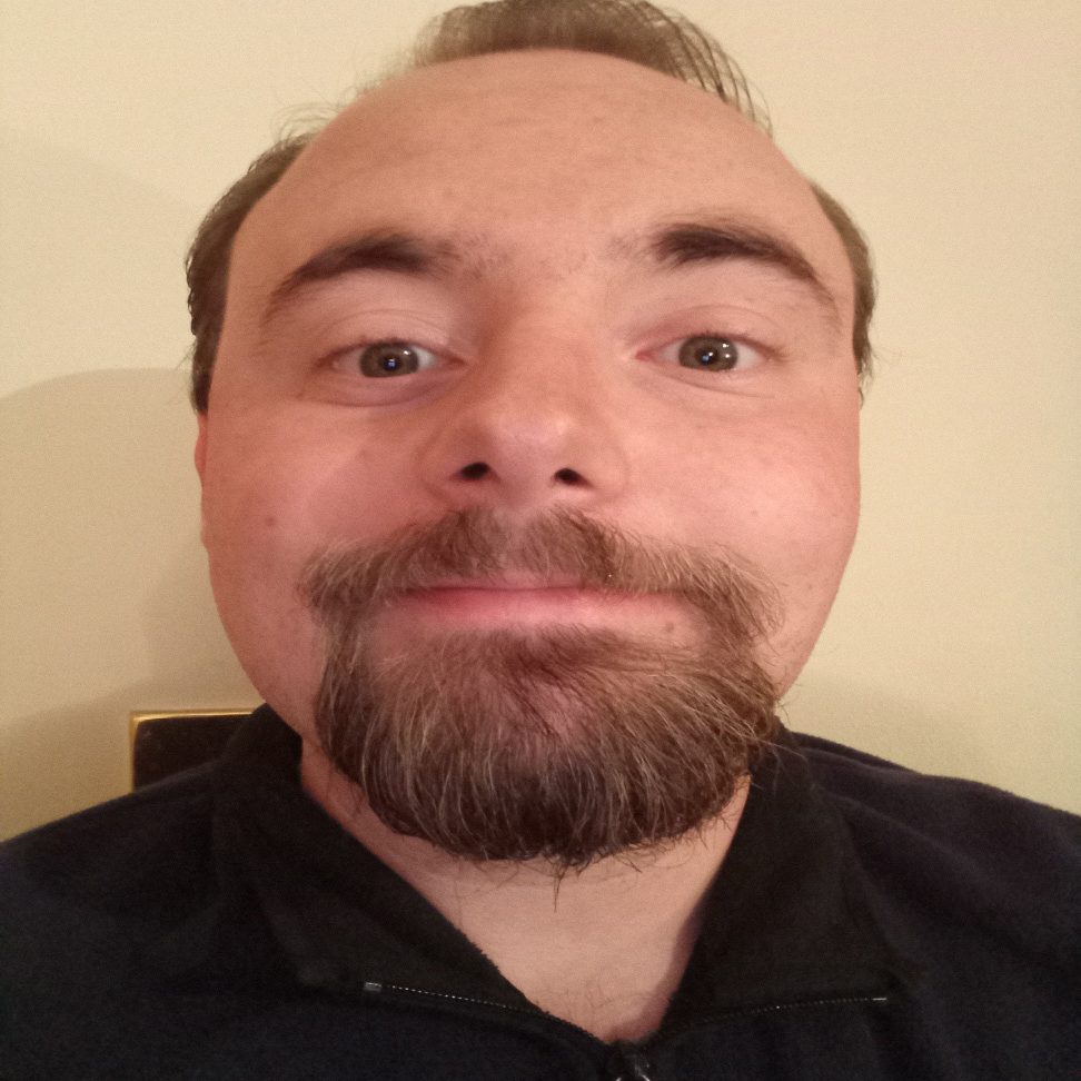 A close-up of a smiling man with a beard, wearing a dark top.