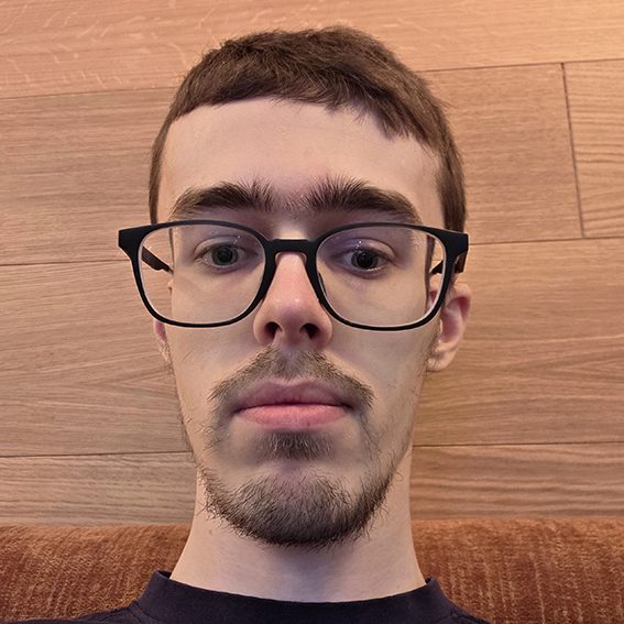 Young man with glasses, short brown hair, and a neutral expression, against a wood background.