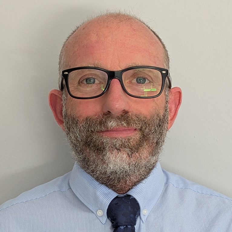 Man with glasses and a beard, wearing a light blue shirt and tie, smiling at the camera.