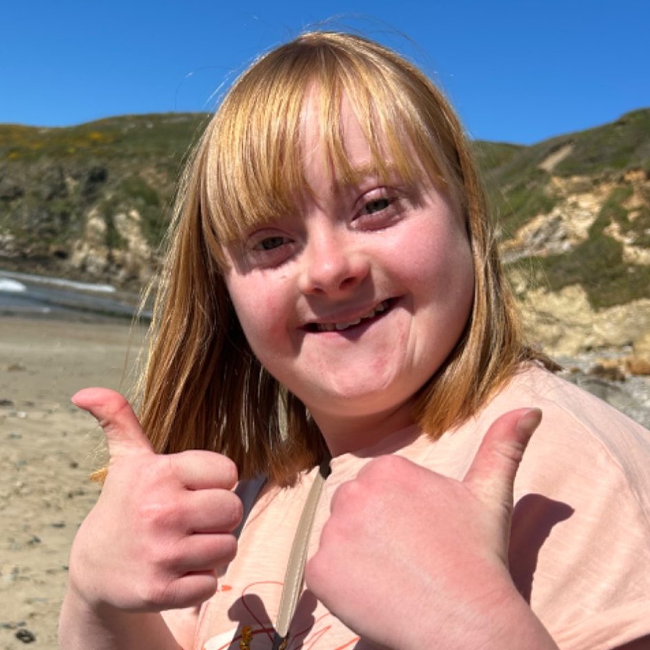 Girl giving a thumbs-up on a beach, smiling with a scenic backdrop.