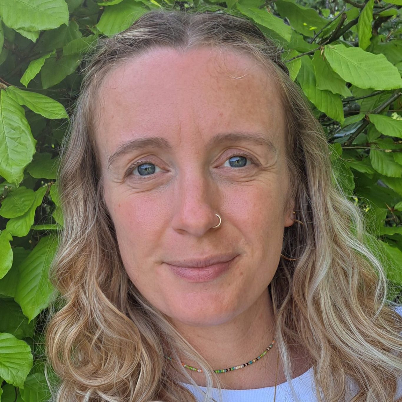 A woman with long wavy hair and a nose ring, smiling in front of green leaves.