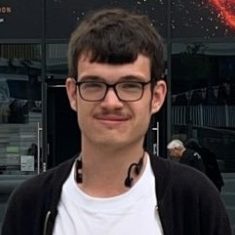 Young man with glasses, light brown hair, and a slight smile, standing outside a building.