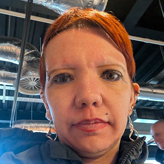 Woman with red hair, wearing earrings, indoors with industrial decor in the background.