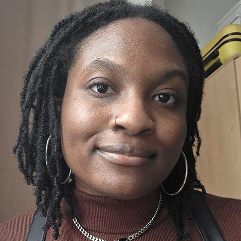Smiling person with braided hair, wearing a brown top and hoop earrings, against a light background.