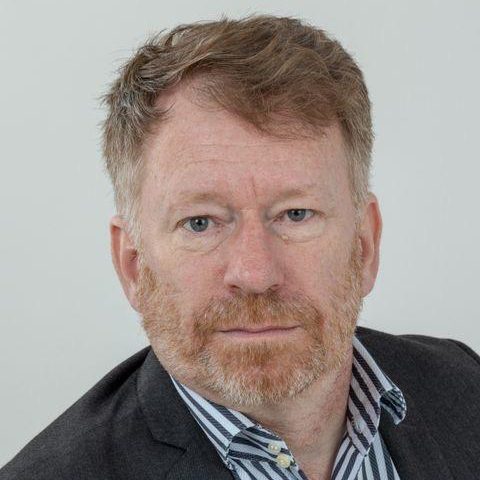 A man with ginger hair and a beard, wearing a grey blazer and striped shirt.