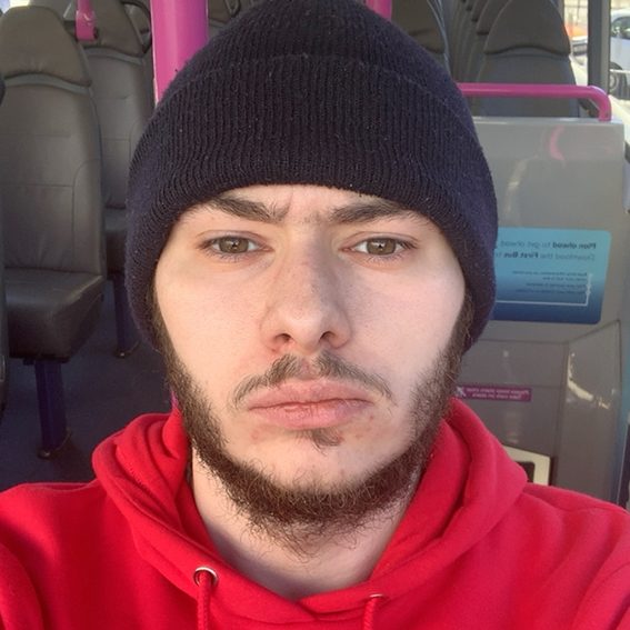Young man with a beard and wearing a black beanie and red hoodie, sitting on a bus.